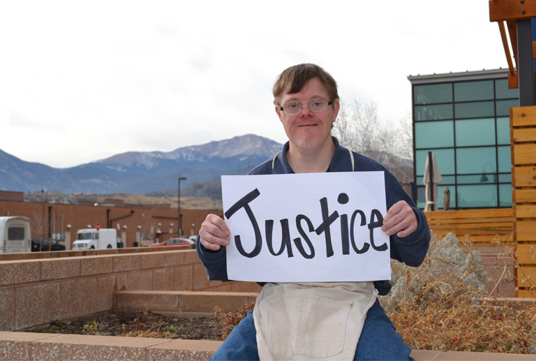 Image of a man with Downs Syndrome holding a sign that reads 'justice' in front of a building and mountains.