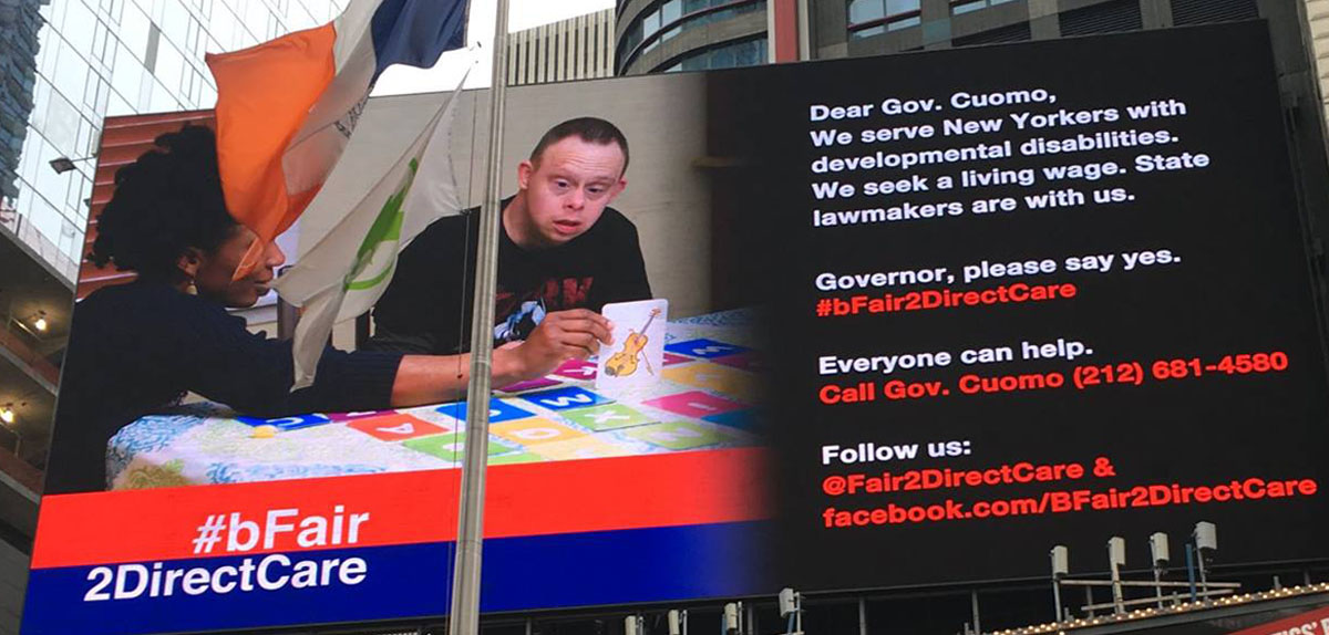 This is a photo of the campaign billboard in Times Square in New York for the bFair2DirectCare campaign. The billboard has a photo of a man with an intellectual/developmental disability at a table with a DSP, and they’re engaged in a literacy task. To the right of the photo on the billboard are these words: Dear Gov. Cuomo, We serve New Yorkers with developmental disabilities. We seek a living wage. State lawmakers are with us. Governor, please say yes. #bFair2DirectCare. Everyone can help. Call Gov. Cuomo (212) 681-458_. Follow us: @Fair2DirectCare & facebook.com/BFair2DirectCare.