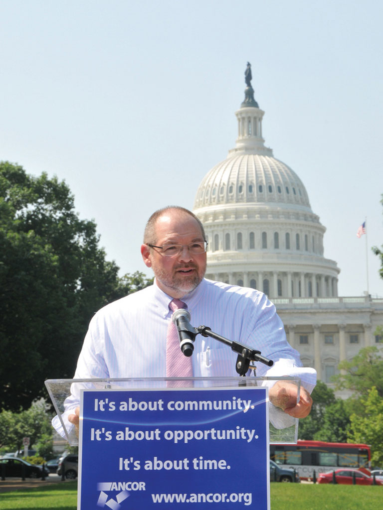 This photo is at the bottom of the second page for the article We Have to Use Every Creative Idea We Have: An Agency Director Looks at the Direct Support Crisis.  The photo shows the article’s author, Christopher Sparks, standing at a podium in front of the U.S. Capitol, speaking at a rally. On the front of the podium is a printed sign that says, It’s about community. It’s about opportunity. It’s about time. ANCOR. www.ancor.org.