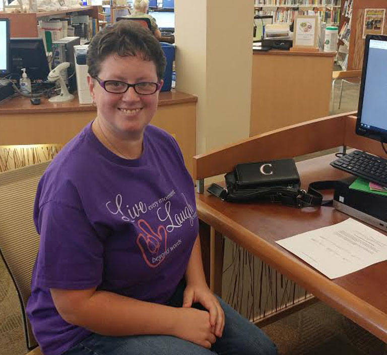 A Photo of Crytsal Arbaugh. She is sitting at a computer at a public library. She is looking at the camera and smiling.