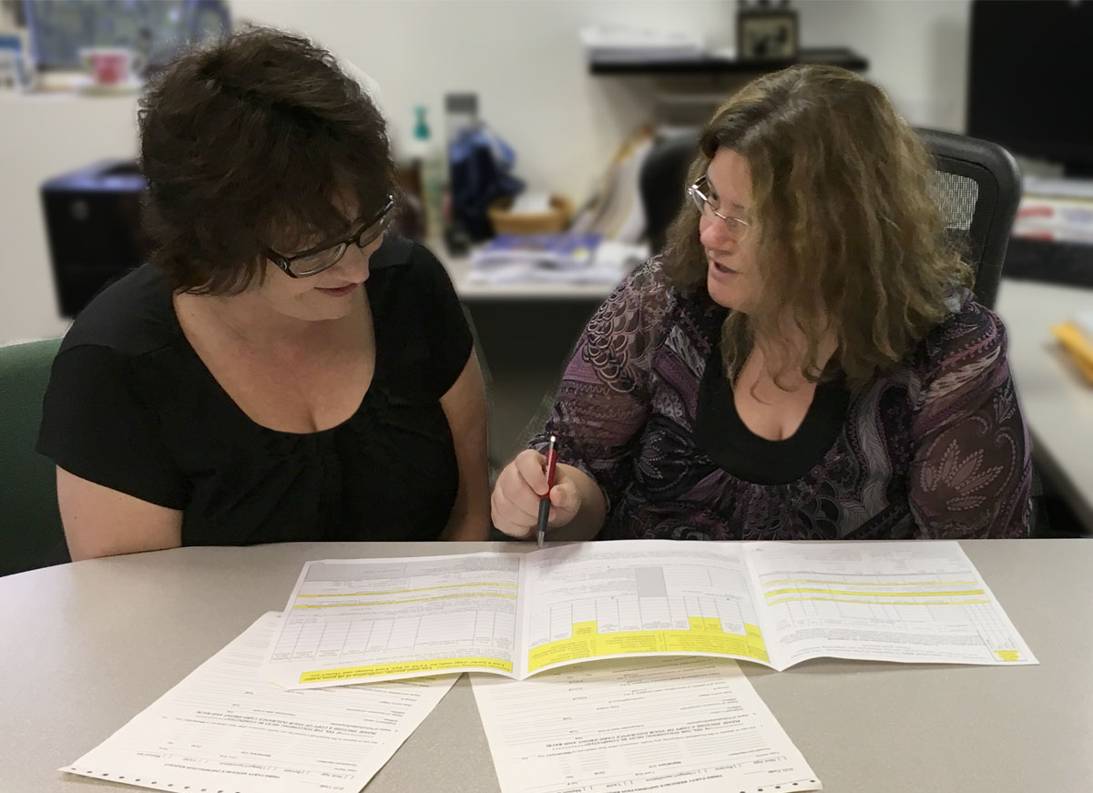 This photo shows the author, Gina Russell, who is a DSP with OHI, sitting at a table with OHI’s Human Resources Manager, Twila Illingworth Tomlin. They are looking at documents on the table in front of them and talking.