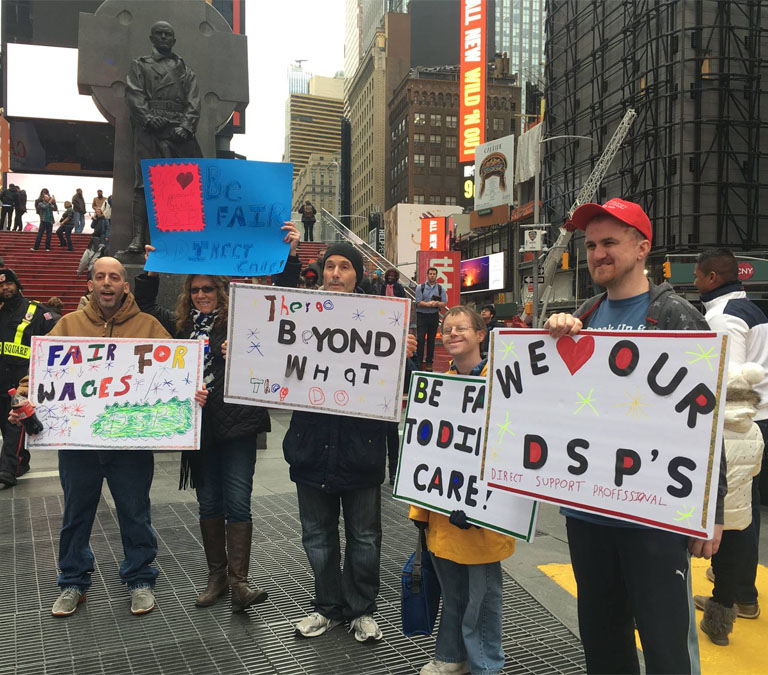This is a photo of five demonstrators with disabilities and DSPs holding signs in Times Square in New York. The signs say We love our DSPs, Be Fair to Direct Care, and two that aren’t fully readable.