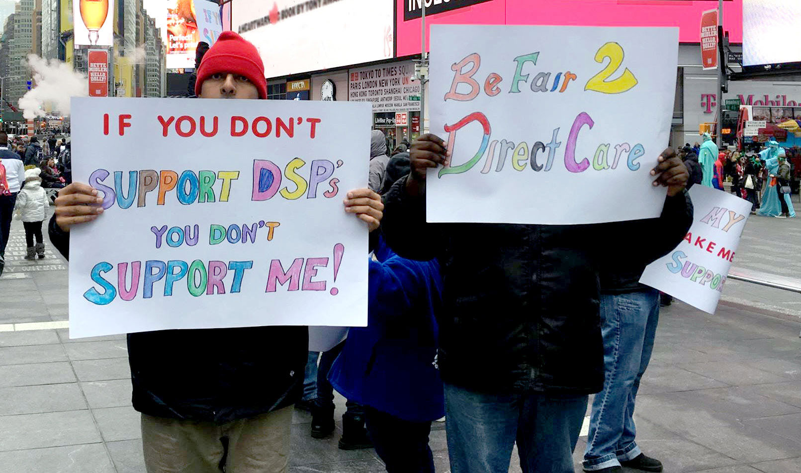 Photo at the top of the opening page for the article "The Direct Support Workforce Crisis: A Systemic Failure." The photo shows two individuals standing in Times Square in New York holding up signs as part of a BFair2DirectCare rally. The signs say, "If you don’t support DSPs you don’t support me!" and "Be Fair 2 Direct Care."