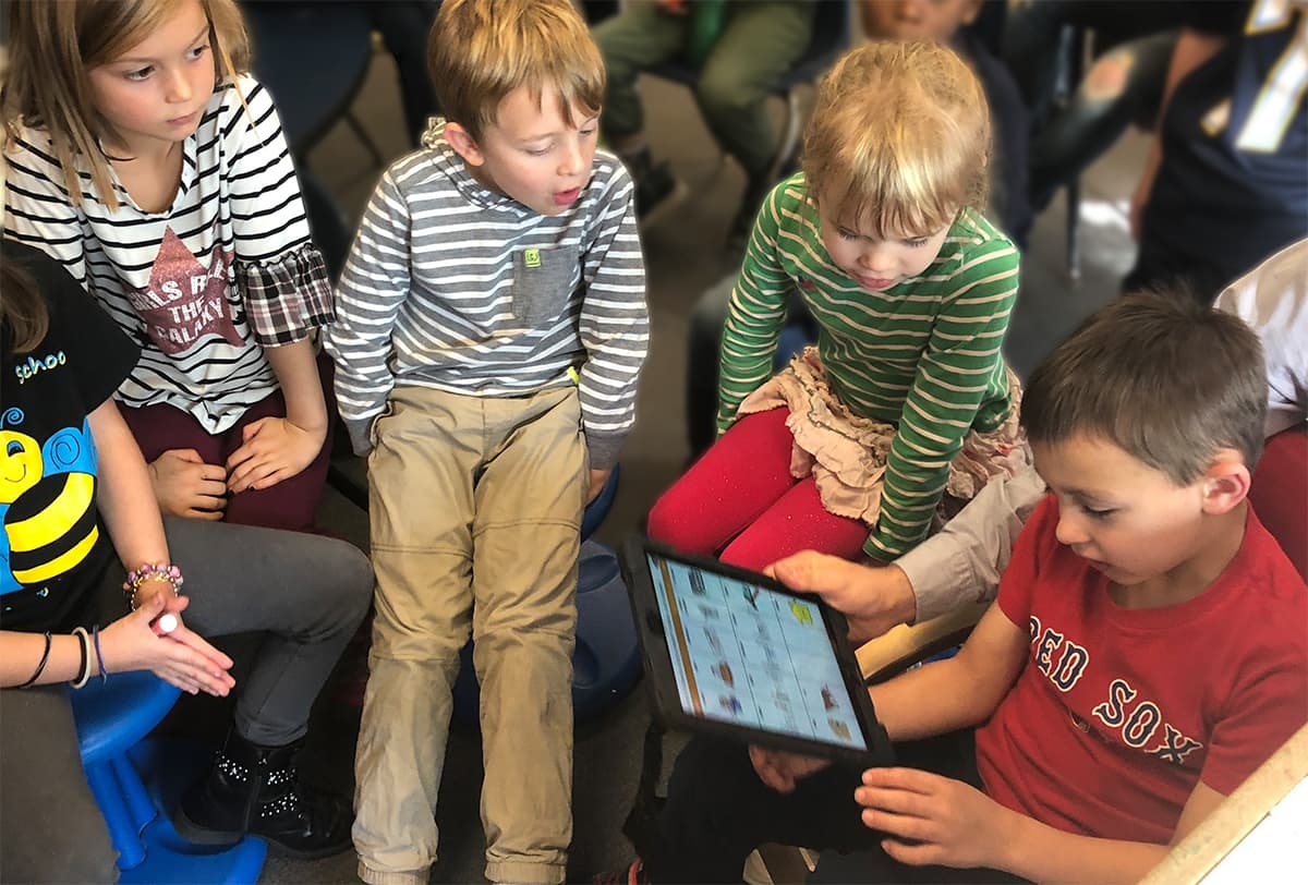 The photo shows four second grade students sitting in chairs in a semi-circle in the classroom, two boys and two girls, and the three students on the left of the photo are looking at the student, Landon Lutz, on the right who’s holding his iPad-like device, which he’s using to select and play greetings for the others.