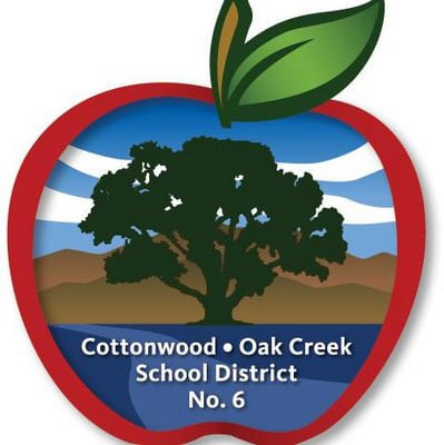 School logo for the Cottonwood-Oak Creek School District. It’s a brightly colored drawing of a big red apple, and within the apple is an Oak tree silhouette, sand-colored mountains in the background, purple landscape in front of the tree, and blue sky with clouds. The district’s name also appears in the apple in white letters.