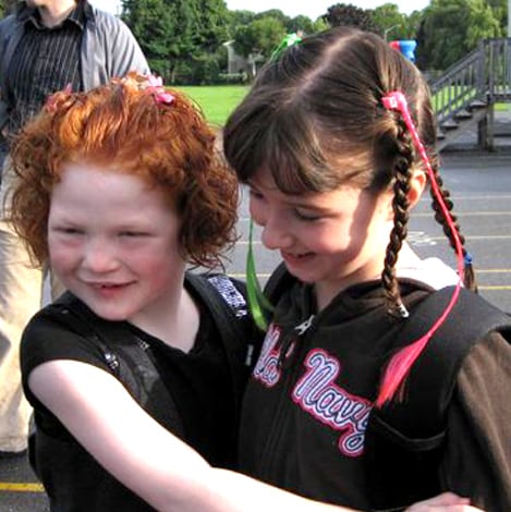 A close-up of the two girls, Maggie and Jordyn, on another occasion standing close to each other in front of the school. They are facing each other and Maggie is hugging Jordyn and they’re smiling.