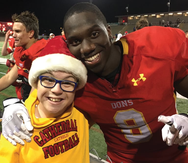 Thomas Byrne, a high school freshman, dressed in his gold Cathedral High School football hooded sweatshirt and huddled at the sideline of the school’s football stadium with his peer mentor and friend, Jordan Genmark Heath, who is in his red football uniform and has just finished a game. Cathedral won the 2016 championship game, and both young men are all smiles.