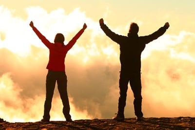 Two people on the top of a mountain. Both people have with their hands up in the air in celebration of their accomplishment.